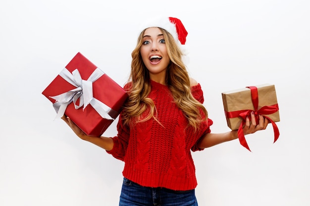 Heureuse femme blonde insouciante célébrant la fête du nouvel an tenant des cadeaux. Porter un bonnet de Noel rouge et un pull en tricot. Poser