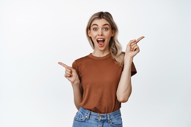 Heureuse femme blonde pointant les doigts sur le côté et souriant, montrant deux options de vente, différentes variantes, debout en t-shirt sur blanc