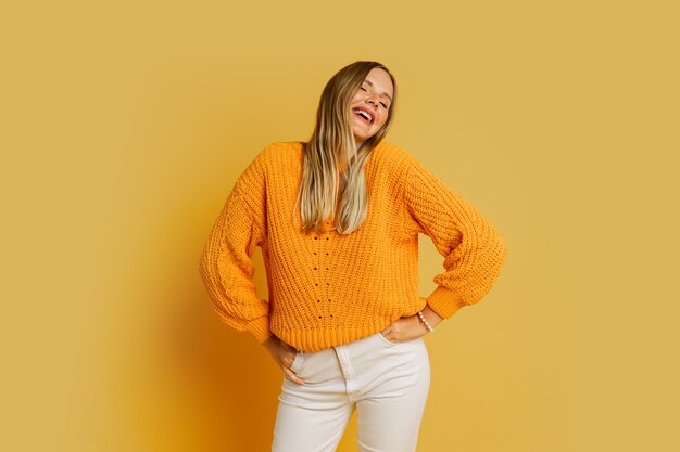 Heureuse femme blonde en pull d'automne élégant orange posant sur jaune.