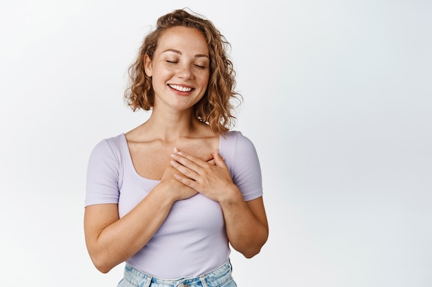 Heureuse femme blonde tenant les mains sur le coeur et souriant, ferme les yeux pensif, rêvant de qch romantique, rêvassant, debout sur blanc.