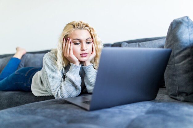 Heureuse femme blonde travaillant sur un ordinateur portable assis sur le canapé dans la maison.