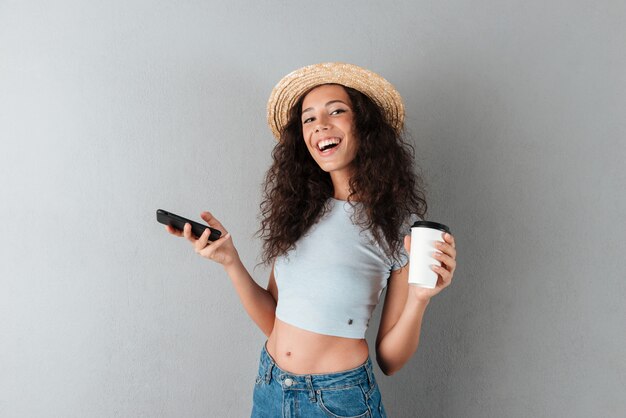 Heureuse femme bouclée au chapeau tenant le smartphone et la tasse de café