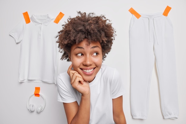 Photo gratuite heureuse femme bouclée curieuse regarde ailleurs les sourires porte avec plaisir un t-shirt décontracté se sent satisfait isolé sur fond blanc avec un pantalon en t-shirt plâtré et un casque remarque quelque chose de bien