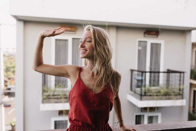 Heureuse femme bronzée en pyjama, agitant la main avec le sourire. Incroyable modèle féminin caucasien debout au balcon.