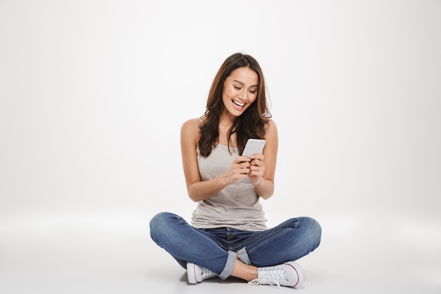 Heureuse femme brune assise sur le sol et écrit un message sur smartphone sur gris