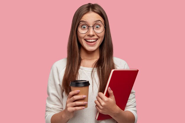 Heureuse femme brune avec une expression excitée, regarde avec bonheur, porte des lunettes rondes, tient un manuel rouge et sort du café, réagit aux bonnes nouvelles de camarade de classe, isolé sur un mur rose