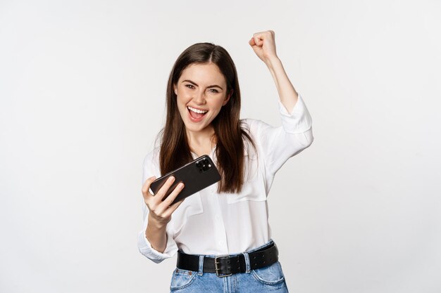 Heureuse femme brune jouant au jeu vidéo mobile, souriant et regardant l'écran excité, debout sur fond blanc