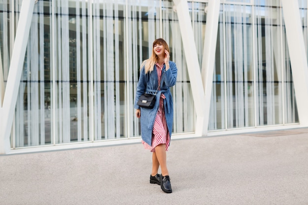 Heureuse femme brune en manteau bleu et robe rouge marchant le long de la rue moderne