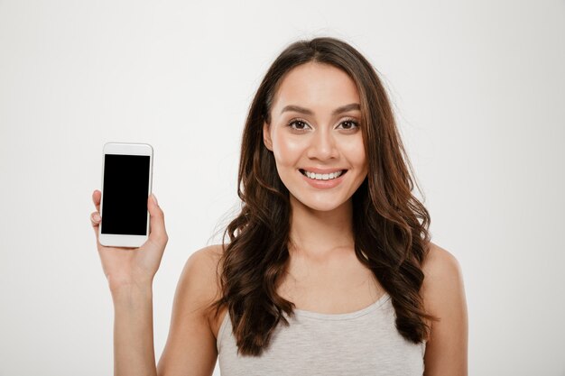 Heureuse femme brune montrant l'écran du smartphone vierge et regardant la caméra sur gris