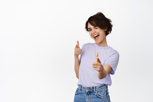 Heureuse femme brune pointant du doigt des pistolets à la caméra, un clin de œil et souriant, fond blanc.