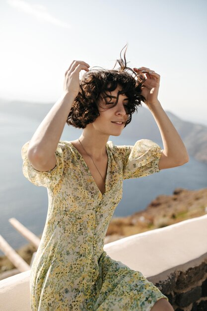 Heureuse femme brune en robe d'été élégante profite d'un temps venteux ensoleillé à l'extérieur Dame en tenue florale est assise sur fond de mer et de montagne et sourit