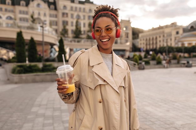 Heureuse femme brune en trench-coat beige surdimensionné écoute de la musique dans des écouteurs rouges, sourit, tient un verre de jus d'orange à l'extérieur