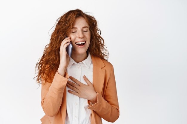 Heureuse femme de bureau rousse parlant sur un téléphone portable et riant d'avoir une conversation décontractée sur un smartphone debout sur fond blanc