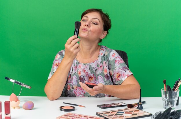 Heureuse femme caucasienne adulte assise à table avec des outils de maquillage tenant du blush et soufflant sur un pinceau de maquillage isolé sur un mur vert avec espace de copie