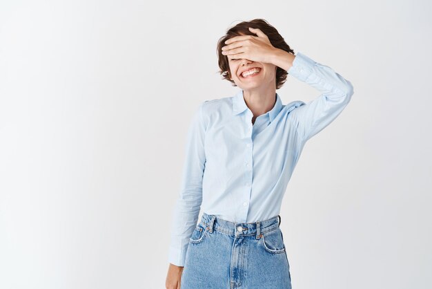 Heureuse femme caucasienne en vêtements de bureau couvrir les yeux avec la main et souriant en attente de surprise debout sur fond blanc