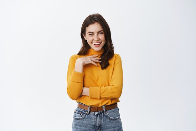 Heureuse femme confiante riant, touchant la poitrine et se montrant un geste d'auto-appréciation, debout dans un pull jaune contre un mur blanc