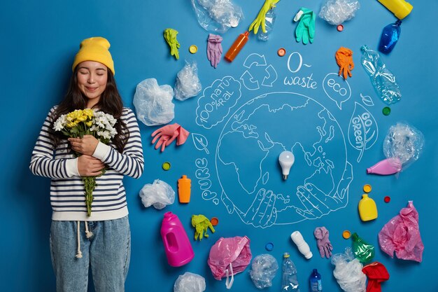 Heureuse femme coréenne satisfaite d'obtenir un bouquet, tient des fleurs blanches et jaunes, se tient contre une planète dessinée et des déchets en plastique sur le mur bleu, nettoie la nature de la pollution.