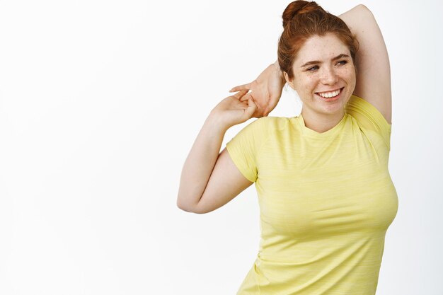 Heureuse femme curvy souriante portant des vêtements de fitness étirant les bras derrière le dos se réchauffer dans la salle de gym debout sur fond blanc