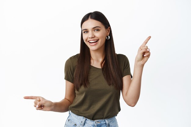 Heureuse femme dansante pointant les doigts sur le côté, souriante et montrant des offres promotionnelles gauche droite, donnant des variantes, montrant une variété de choix, mur blanc