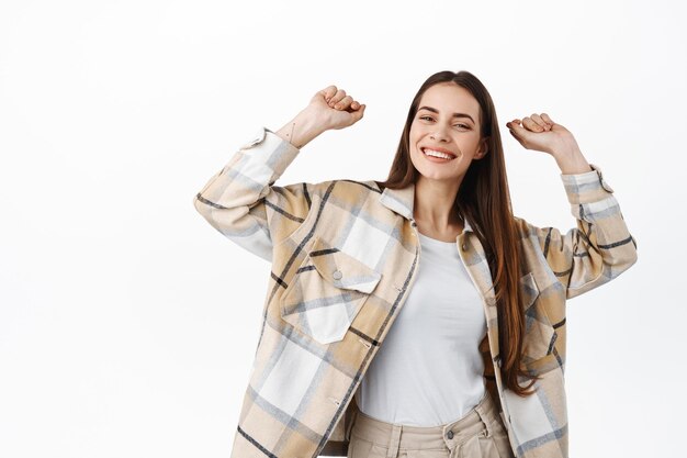 Heureuse femme dansante s'amusant. Une fille élégante danse les mains en l'air et sourit, célèbre quelque chose, se dresse sur fond blanc en chemise à carreaux