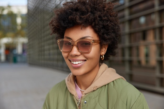 Heureuse femme élégante aux cheveux bouclés porte une veste de lunettes de soleil à la mode et un sweat à capuche sourit joyeusement à l'extérieur des poses sur fond flou profite du temps libre Concept de style de vie et de style des gens