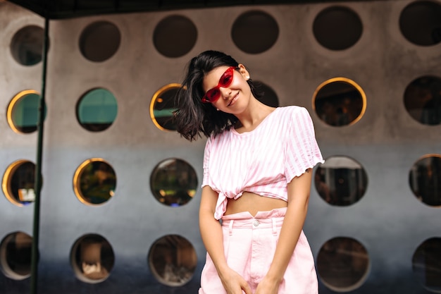 Heureuse femme élégante regardant la caméra sur fond urbain. Tir extérieur d'une femme magnifique à lunettes de soleil roses posant avec le sourire.