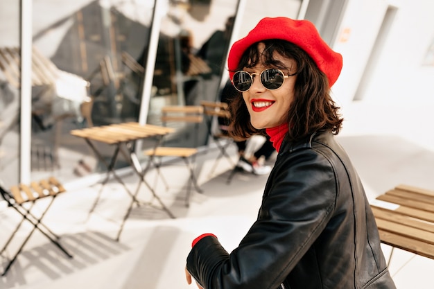 Photo gratuite heureuse femme élégante en tenue française avec des lèvres rouges et béret rouge assis en terrasse extérieure