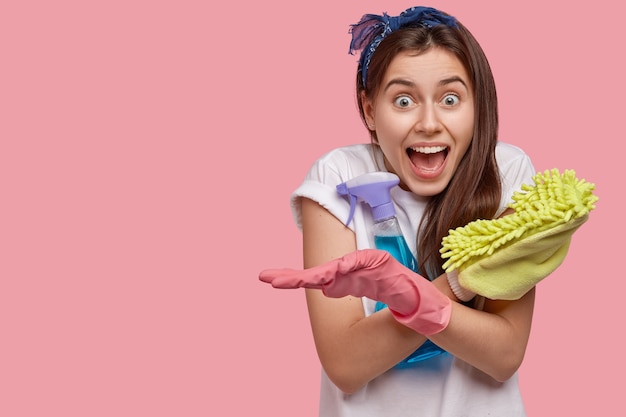 Heureuse Femme émotionnelle Croise Les Mains, Tient Une Vadrouille Et Un Nettoyant En Aérosol, Porte Un T-shirt Et Des Gants Blancs, Heureuse De Terminer Les Travaux Ménagers à Temps, Pas En Retard Pour La Date, Pose Contre Le Mur Rose. Bonne Humeur Pour Le Nettoyage