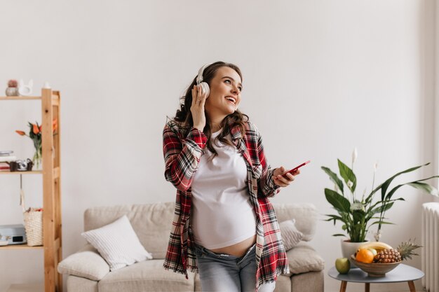 Heureuse femme enceinte brune en pantalon denim et tee-shirt blanc, écouter de la musique dans les écouteurs