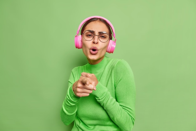 Heureuse femme enjouée chante la chanson indique que vous appréciez la musique préférée dans des écouteurs roses apprécie le moment porte des lunettes transparentes et des poses de col roulé contre le mur vert