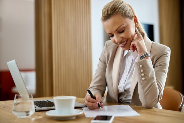 Heureuse femme entrepreneur prenant des notes tout en travaillant sur un ordinateur portable dans une chambre d'hôtel