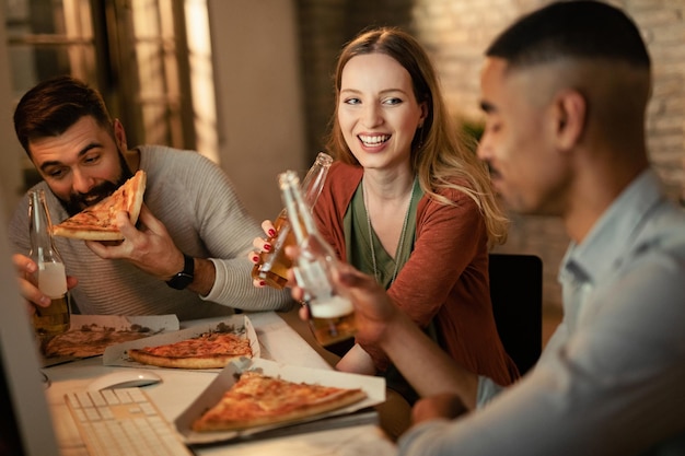 Heureuse femme entrepreneure et ses collègues masculins mangeant de la pizza et buvant de la bière pendant une pause au bureau