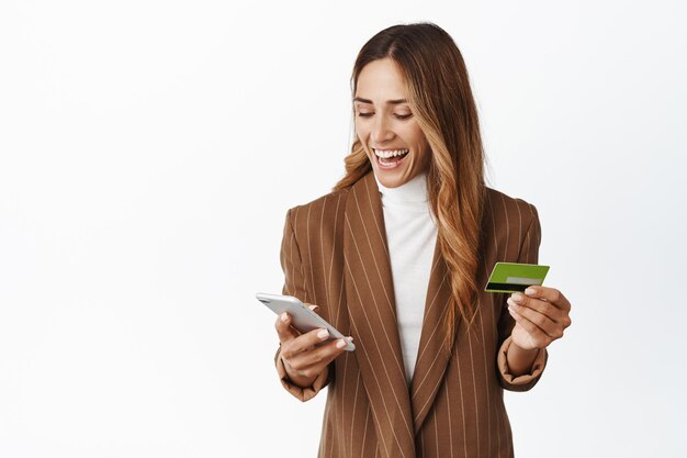 Heureuse femme d'entreprise commande en ligne sur l'application pour smartphone détenant une carte de crédit et souriant sur fond blanc de téléphone portable