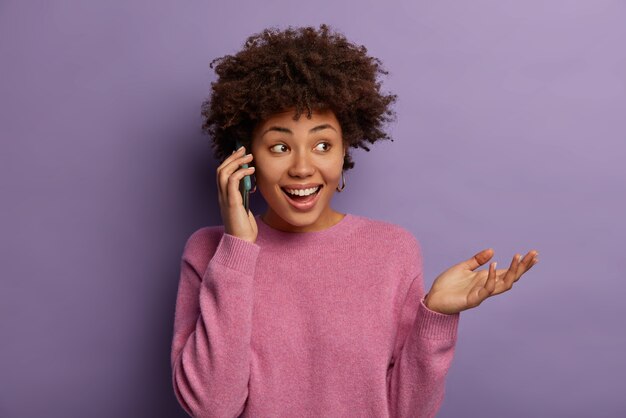 Heureuse femme ethnique parle au téléphone, regarde ailleurs et fait des gestes, discute avec impression de sa visite au théâtre, vêtue d'un pull décontracté, concentrée de côté, a un sourire à pleines dents isolé sur un mur violet