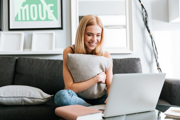 Heureuse femme étreignant oreiller et utilisant un ordinateur portable sur le canapé