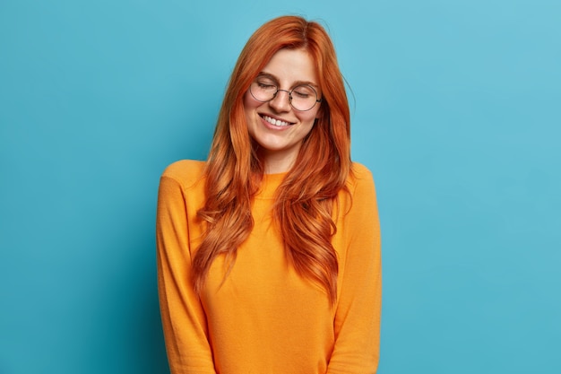 Heureuse femme européenne au gingembre aux cheveux longs garde les yeux fermés et sourit, porte agréablement de grandes lunettes rondes et un pull orange.