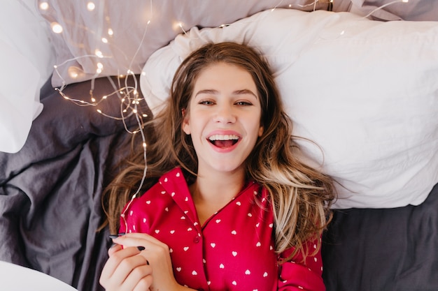 Heureuse femme européenne en pyjama rouge allongé sur un drap noir. Photo intérieure d'une fille aux cheveux noirs émotionnelle riant dans son lit.