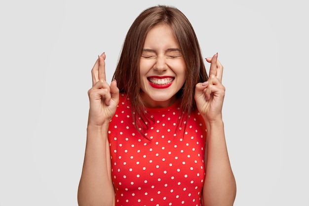 Heureuse femme européenne séduisante fait le souhait de gagner, lève les mains avec les doigts croisés, attend les résultats de la loterie, ferme les yeux