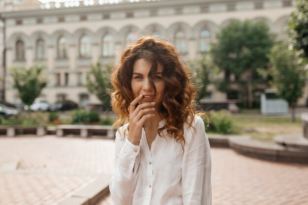 Heureuse femme excitée avec des boucles touchant ses lèvres et posant sur les bâtiments de la vieille ville