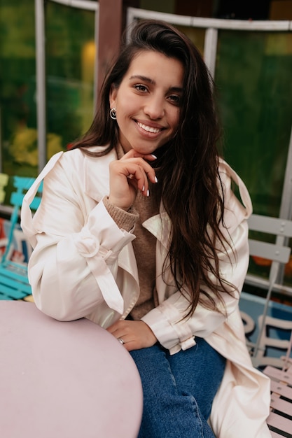 Heureuse femme excitée avec de longs cheveux noirs portant un manteau beige assis dans une cafétéria de l'espace ouvert et en attente de café