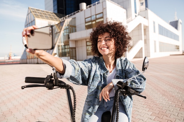 Heureuse femme frisée assise sur une moto moderne à l'extérieur