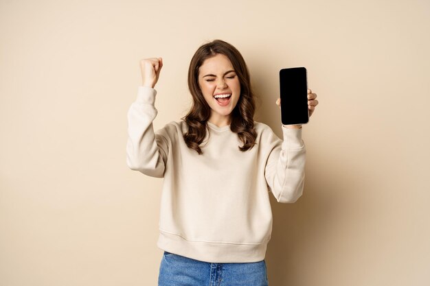 Heureuse femme gagnante montrant l'écran du smartphone et criant de joie pompe à poing debout sur le dos beige...