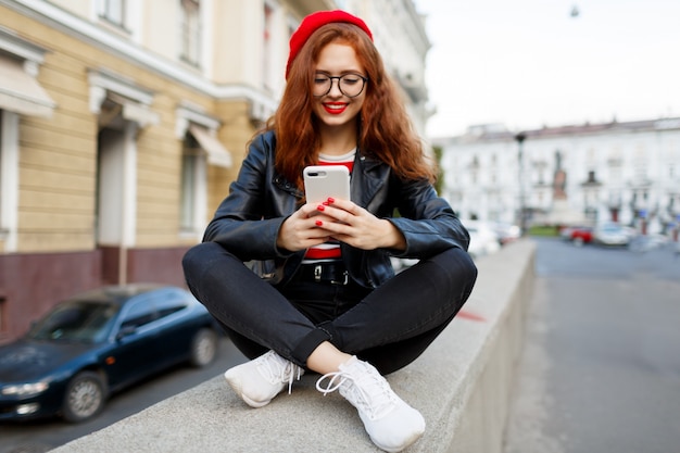 Heureuse femme gingembre fabuleux en élégant béret rouge dans la rue à l'aide de smartphone