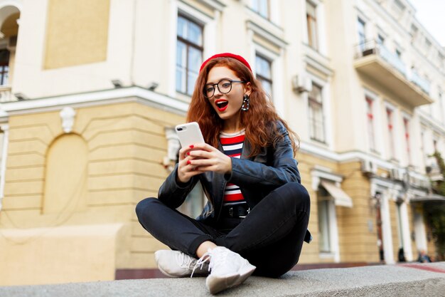 Heureuse femme gingembre fabuleux en élégant béret rouge dans la rue à l'aide de son smartphone