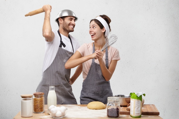 Heureuse femme et homme stupide à la cuisine, se battre avec un fouet et un rouleau à pâtisserie, avoir des expressions joyeuses