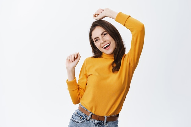 Heureuse femme libre s'amusant, dansant les mains en l'air et regardant l'espace vide, debout sur du blanc dans des vêtements décontractés