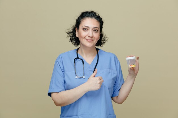 Heureuse femme médecin d'âge moyen portant un uniforme et un stéthoscope autour du cou montrant un récipient de mesure avec des pilules dedans regardant la caméra montrant le pouce vers le haut isolé sur fond olive