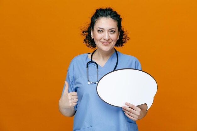 Heureuse femme médecin d'âge moyen portant un uniforme et un stéthoscope autour du cou regardant la caméra montrant une bulle de chat et le pouce vers le haut isolé sur fond orange