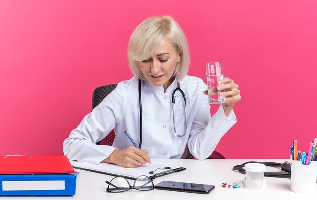 Heureuse femme médecin slave adulte en robe médicale avec stéthoscope assis au bureau avec des outils de bureau tenant un verre d'eau et écrivant sur un presse-papiers isolé sur fond rose avec espace de copie