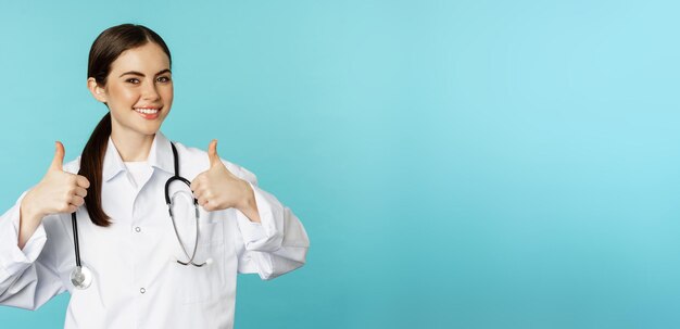Heureuse femme médecin travailleur médical en blouse blanche montrant les pouces vers le haut dans l'approbation comme quelque chose de louange